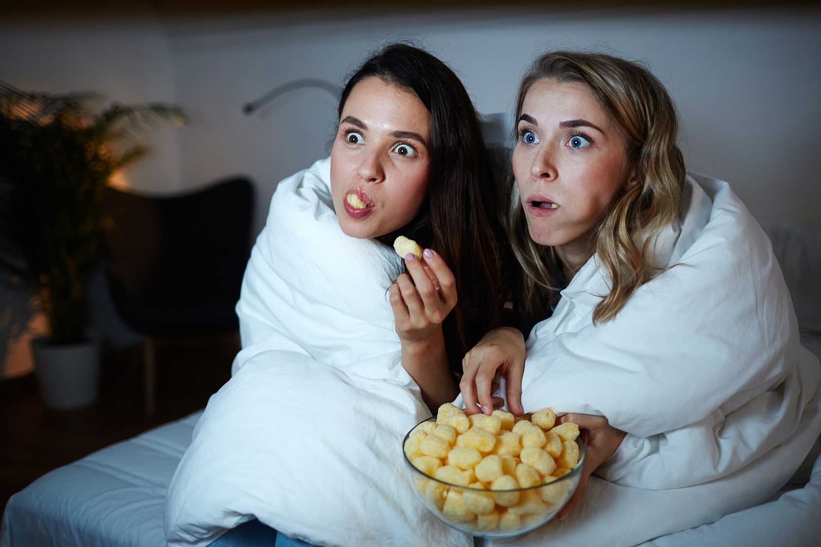 Curious girls eating corn rolls from bowl while watching captivating movie at night